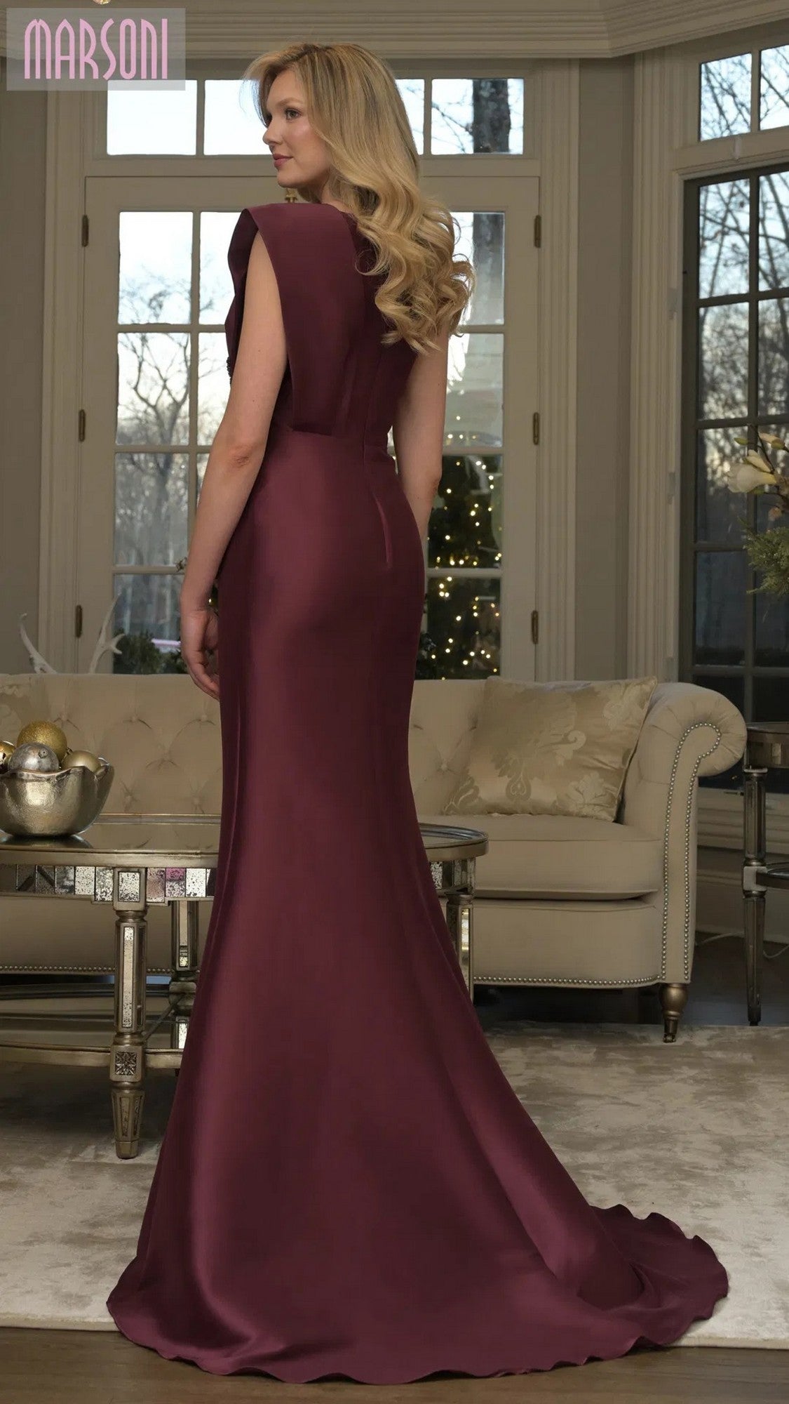A woman in the Marsoni MV1359 long burgundy sleeveless formal dress with a mermaid skirt stands in a living room featuring large windows, a beige sofa, and a decorative table.
